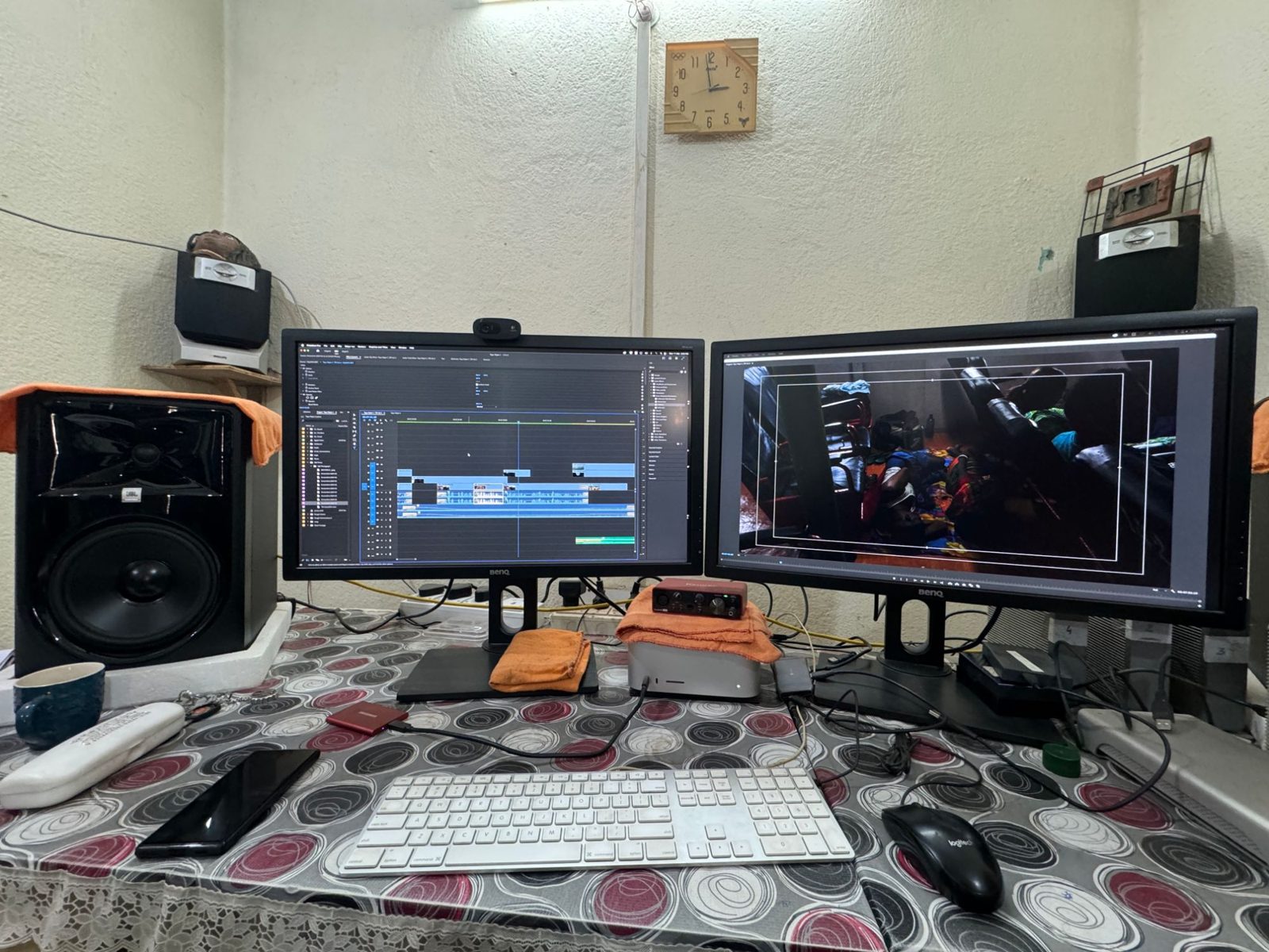 An editing desk with two monitors, one showing a video timeline and the other a dim interior shot, between studio speakers
